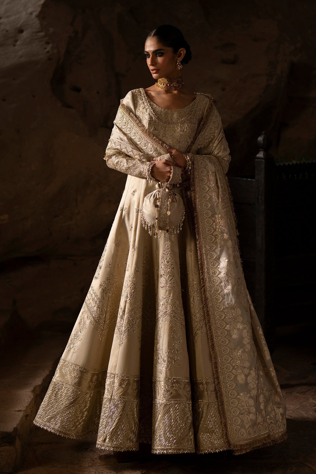 Woman in an ornate beige dress standing against a textured wall.